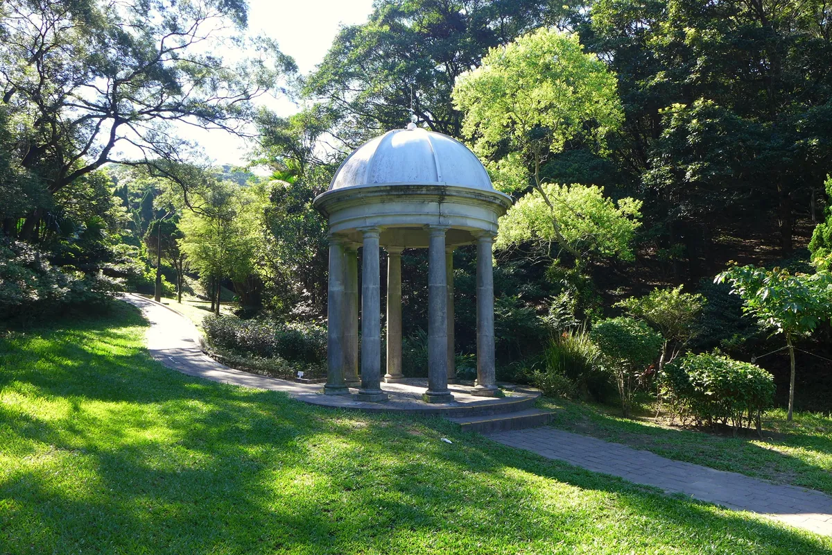 A stone pavilion with a domed roof on a lawn in Victoria Peak Garden, surrounded by trees