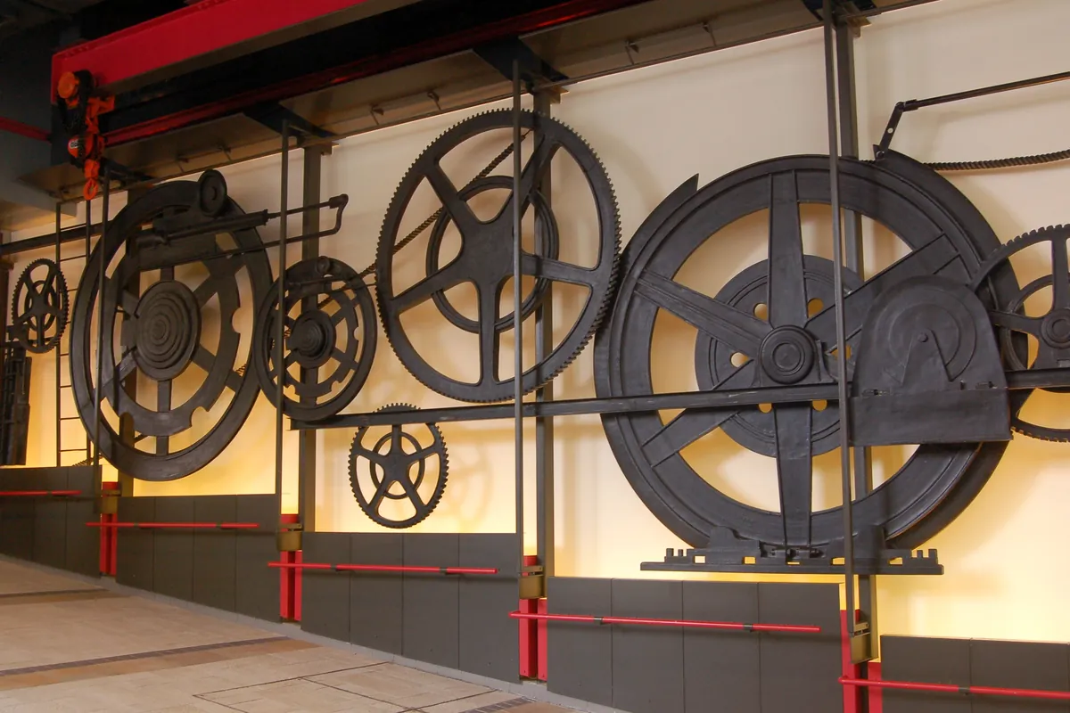 Large black iron gear wheels mounted on a cream wall at the upper station of the Peak Tram, lit from below