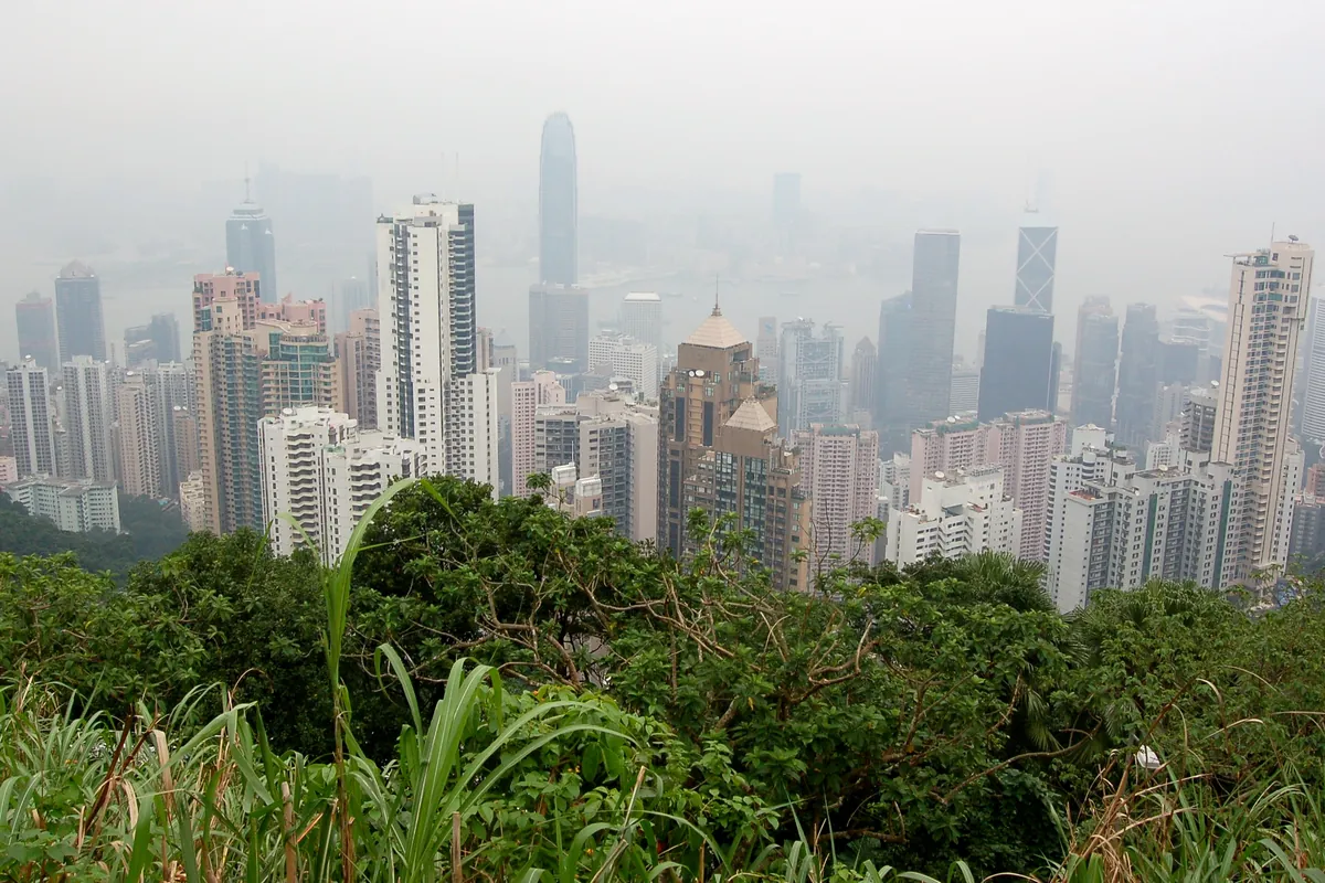 Hong Kong skyscrapers fading into pale haze above a green hillside, seen from the Peak Tower